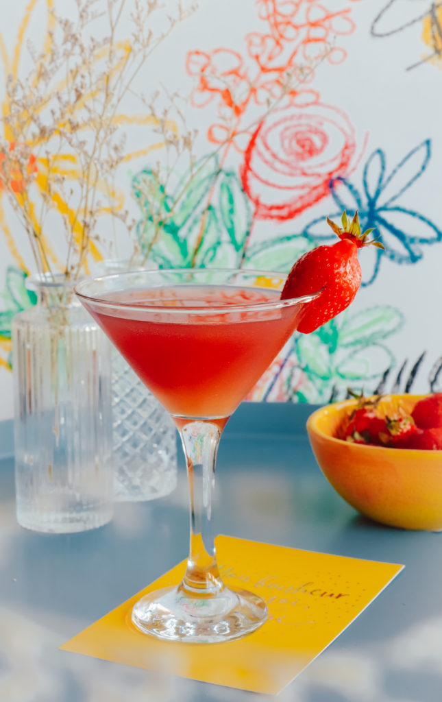 Klart cocktailglass stående på et gult kort med grafitti i bakgrunnen. Glasset inneholder rød daquari og har et jordbær på kanten av glasset.