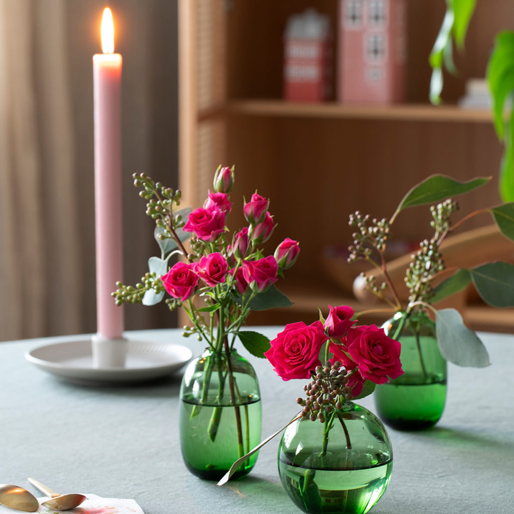 Tre små grønne glassvaser med mørk rosa roser og grønne planter, med et lyserosa stearinslys i bakgrunnen