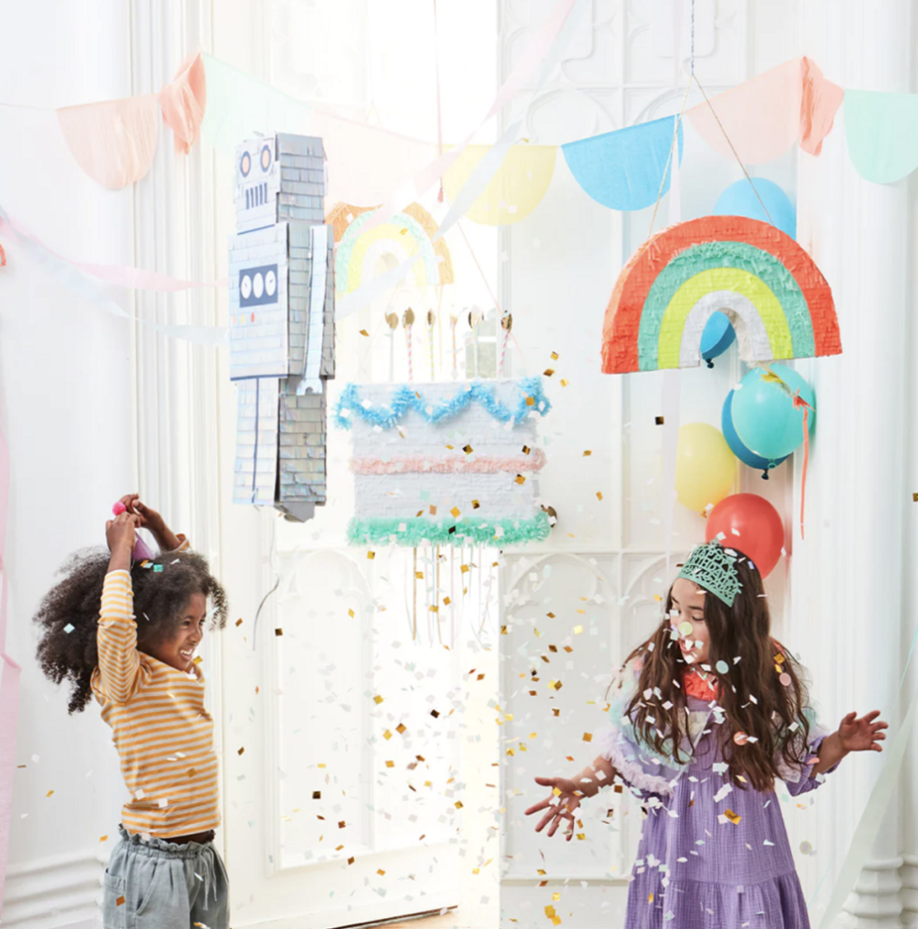 To barn i en bursdagsfest som står under 3 stk ulike piñata, hvor konfetti daler ned.